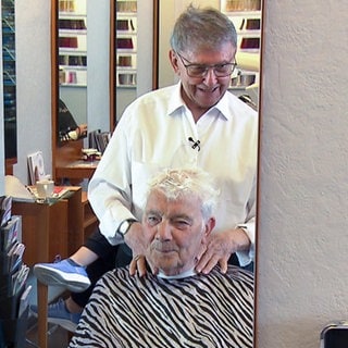 Günter ist 90 Jahre alt und Friseurmeister in Rente. Für seine Freunde und Bekannten kommt er aber gerne aus dem Ruhestand zurück. Günter steht hinter einem Kunden am Friseurstuhl. 