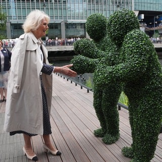 Königin Camilla streckt die Hand zwei Moos-Menschen entgegen. Sie lacht dabei und trägt einen hellen Trenchcoat.